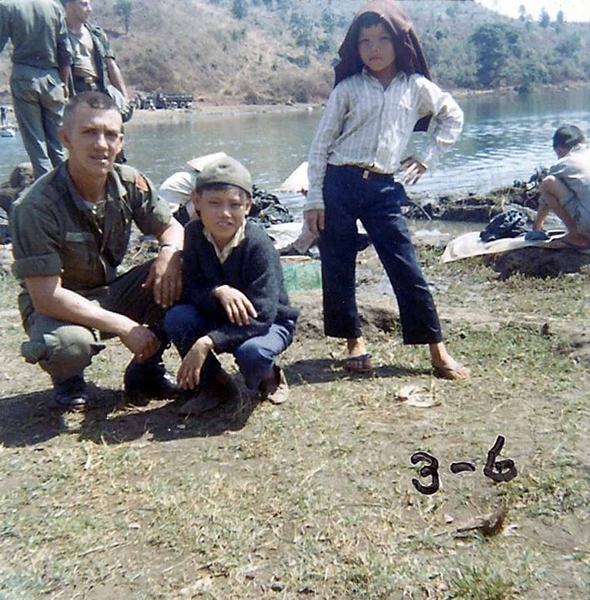 Fresh Haircut
Photo taken at Bien Ho, a lake near our base camp at Pleiku. This is the lake where we did laundry alongside the Vietnamese. 
Sgt Joe Cook poses with two Vietnamese children.  The boy next to Joe spoke English; his name is "Co" and he is wearing Joe's hat (backwards, naturally).  The boy in the white shirt is Con Tin. Picture taken before our 'B" Battery ever went to the field to stay. Date, approximately January 1966. Joe is sporting a new haircut from a Vietnamese barber.
